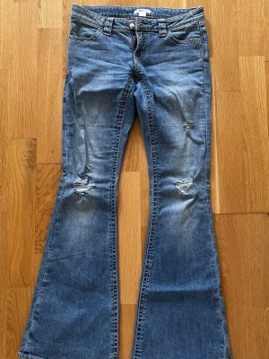 Blå bootcut jeans med slitningar - Säljer ett par blå bootcut jeans med slitningar på knäna och broderade detaljer på bakfickorna. Jeansen har låg midja och klassisk femficksmodell. Materialet är denim i bomull med lite stretch för skön passform.