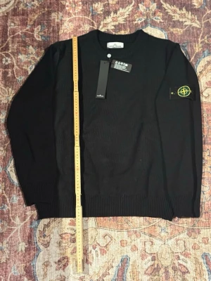 Svart stickad tröja från Stone Island - Snygg svart stickad tröja från Stone Island med klassisk rund hals och ribbade muddar. Tröjan har den ikoniska Stone Island-loggan på vänster ärm och är tillverkad i ett mjukt, tjockt material som passar perfekt till höst och vinter.