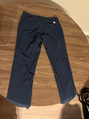 Blå golfbyxor från Adidas - Mörkblå golfbyxor från Adidas med diskret logga på vänster höft. Byxorna har raka ben och elastisk midja. Tillverkade i ett lätt och stretchigt material som ger bra rörelsefrihet.