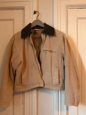 Beige kort jacka med mörk krage - Snygg beige jacka i kort modell med mörkbrun krage. Jackan har dragkedja framtill, en bröstficka med dragkedja och två sidofickor. Tillverkad i ett robust bomullstyg, perfekt för lager på lager under kyligare dagar.
