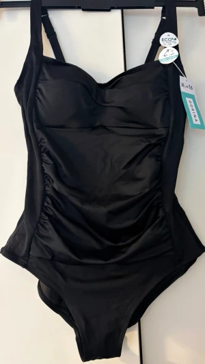 Svart baddräkt från Dorina - Stilren svart baddräkt från Dorina med breda axelband och lätt rynkade sidor för en smickrande passform. ”Tummy control”, shape effekt. Tillverkad i ett mjukt, stretchigt material som känns bekvämt mot huden. Perfekt för strand eller poolhäng. Strl.XL/46/48.