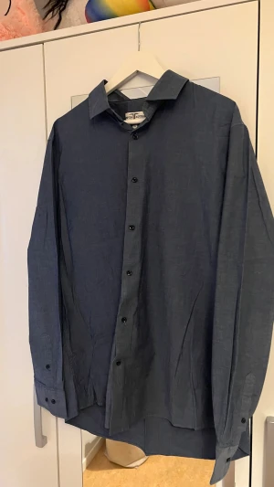Blå skjorta från Hampton Republic XXL - Snygg blå skjorta från Hampton Republic i storlek XXL. Slim fit-modell med comfort stretch för extra bekvämlighet. Klassisk krage, långa ärmar och knäppning framtill. Perfekt för dig som vill ha en stilren och enkel look.