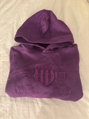 Lila hoodie från Gant med brodyr - Säljer en lila hoodie från Gant med broderad logga och texten 'American Sportswear' framtill. Hoodien har huva, ribbade muddar och är gjord i mjukt material. Hoodien är i storlek 158/164 och är helt ny med prislapp!💕