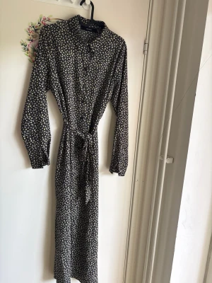 Blommig långklänning från Vero Moda - Långklänning från Vero Moda med svart botten och små blommor i vitt, gult och lila. Klänningen har lång ärm, knappar framtill och ett knytband i midjan. Perfekt för dig som gillar en feminin och avslappnad look.