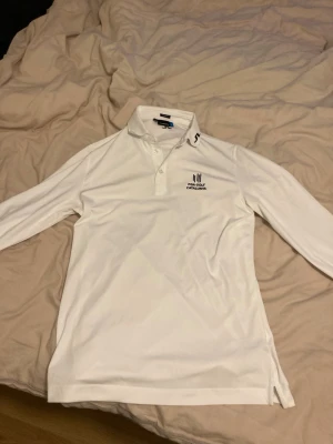 J Lindberg golf overshirt - Säljer en vit långärmad golfpiké av J.Lindberg, broderad logga för PGA Golf Catalunya på bröstet. Tröjan har krage med knappslå och svart detalj på kragen. Perfekt för golfbanan eller sportiga dagar.