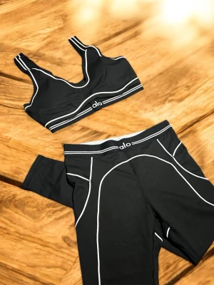 Svart träningsset från Alo - Snyggt svart träningsset från Alo med vita kontrastsömmar. Setet består av en sport-bh med breda axelband och matchande tights med hög midja. Perfekt för dig som vill ha både stil och komfort på gymmet.