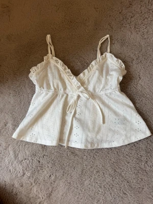 Vit omlottopp med spetsdetaljer - Söt vit omlottopp med tunna axelband och volangkant längs ringningen. Toppen har broderade hål och knyt i midjan för en feminin look. Perfekt för varma dagar och ger en romantisk vibe till din outfit. Storleken är M men den känns väldigt mycket mindre. Hade jag gissat utan att titta på lappen skulle jag gissat på xs eller s. 