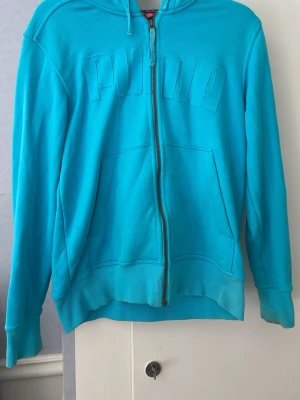 Turkos hoodie med dragkedja från Puma - Snygg turkos hoodie från Puma med dragkedja framtill och stor ficka på magen. Hoodien har huva med dragsko och ribbade muddar vid ärmslut och nederkant. Puma-loggan är broderad ton-i-ton på bröstet. Perfekt för en sportig och avslappnad stil.