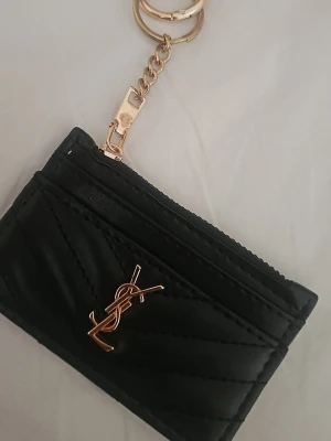 Svart korthållare  - Snygg svart korthållare i quiltat läder med guldfärgad logga framtill. Har flera kortfack och en dragkedja upptill samt en guldfärgad nyckelring. Perfekt accessoar för att hålla koll på kort och nycklar.