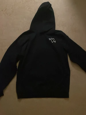 Svart hoodie med hjärta och pil - Säljer en svart hoodie med huva och ett vitt tryck av ett hjärta genomborrat av en pil på bröstet. Hoodien har klassisk passform och ribbade muddar vid ärmslut och nederkant. Perfekt för en chill och avslappnad stil.