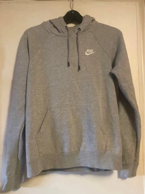 Grå hoodie från Nike med logga - Klassisk grå hoodie från Nike med vit broderad logga på bröstet. Hoodien har dragsko i huvan och en stor magficka framtill. Tillverkad i mjukt sweatshirtmaterial och har ribbade muddar vid ärmar och nederkant. Perfekt för en avslappnad och sportig stil.