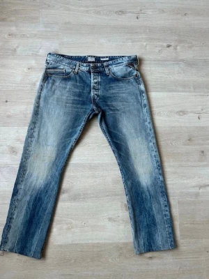 Blå Replay Waitom Jeans med slitningar  - Ett par blå Replay Waitom jeans med raka ben och tydliga slitningar på framsidan. Jeansen har klassisk femficksmodell och ljus tvätt med kontrasterande sömmar. Materialet är denim i bomull och passformen är avslappnad. W38, passar även W36 och kanske W34 om man vill ha jeansen lite baggy