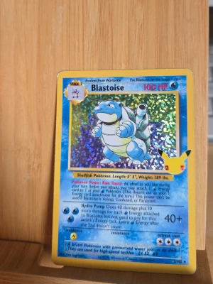 Blastoise Pokémonkort celebrations - Säljer ett holografiskt Blastoise Pokémonkort med gul ram och glittrande bakgrund. Kortet har 100 HP och visar Blastoise i blått och vitt. Baksidan har klassiskt Pokémon-motiv med Pokéboll. Perfekt för samlare eller Pokémonfans!