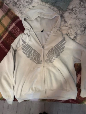 Vit hoodie med vingar framtill - Säljer en vit hoodie med dragkedja och stora gråa vingar tryckta på bröstet. Hoodien har huva med dragsko och två fickor framtill. Perfekt för dig som gillar streetwear och vill sticka ut lite extra. Har en målarfärgs fläck på luvan som itne går bort 