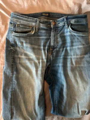 Blå jeans från Jack & Jones - Säljer ett par klassiska blå jeans från Jack & Jones. Jeansen har fem fickor, normal passform och är tillverkade i ett mjukt denimtyg. Snygg tvätt med ljusa partier och subtila slitningar för en avslappnad look.