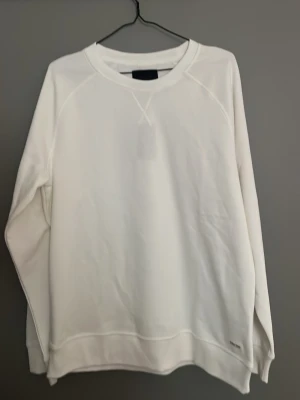 Vit basic sweatshirt - Helt oanvänd stilren vit sweatshirt från Lager157 med rund hals och långa ärmar. Tröjan har en klassisk passform och ribbade muddar vid ärmslut och nederkant. Perfekt för en avslappnad och enkel look.