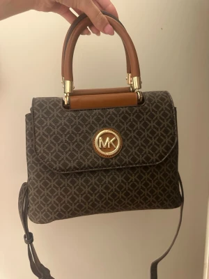 Brun handväska  - Snygg brun handväska med guldfärgade detaljer och logotyp framtill. Väskan har ett geometriskt mönster, ett stadigt handtag och en avtagbar axelrem. Perfekt accessoar för dig som gillar klassisk och trendig design.