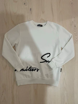 Vit sweatshirt med svart text - Säljer en stilren vit sweatshirt med svart textdetalj nertill på framsidan. Tröjan har rund halsringning, långa ärmar och ribbade muddar vid ärmslut och nederkant. Perfekt för en avslappnad och trendig look.