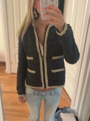 Svart pufferjacka med beige detaljer - Säljer en svart pufferjacka med beige fransdetaljer längs kanter, fickor och ärmslut. Jackan har dragkedja framtill och två fickor på varje sida. Den är quiltad och har en rak, kortare passform. Perfekt för kalla dagar.