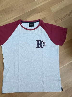 Grå och vinröd t-shirt med tryck - Snygg t-shirt i basebollstil med grå kropp och vinröda ärmar. Tryck med bokstäverna 'R's' på bröstet. Rund halsringning med vinröd kant. Perfekt för en avslappnad look och enkel att matcha med jeans eller shorts.
