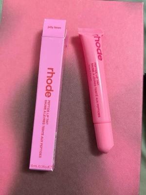 rhode Peptide Lip Tint Jelly Bean - Läppbalsam från rhode i färgen Jelly Bean. Kommer i en rosa tub med matchande förpackning och har en smidig applikator. Innehåller 10 ml och är berikat med peptider för extra vård och glans till läpparna. Perfekt för dig som vill ha återfuktade och snygga läppar.