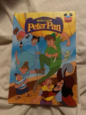 Peter Pan Disney Klassiker bok - En färgglad inbunden bok med Peter Pan från Walt Disney Classics. Omslaget har illustrationer av Peter Pan, Tingeling, Kapten Krok och andra karaktärer. Baksidan är helt gul utan text eller bilder. Perfekt för dig som gillar klassiska äventyr.