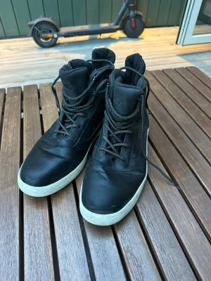 Svarta höga sneakerskängor - Säljer ett par svarta höga sneakerskängor med vita sulor och svarta snören. Kängorna har vadderad insida och detaljer i textil på sidorna. Perfekta för dig som vill ha en sportig och urban look till hösten eller vintern.