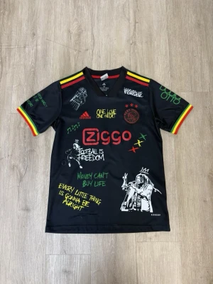 Ajax x Bob Marley svart matchtröja - Snygg svart Ajax matchtröja i samarbete med Bob Marley och Adidas. Tröjan har färgglada detaljer i rött, gult och grönt på axlar och ärmslut, samt tryck med texter och illustrationer inspirerade av reggae och fotboll. Perfekt för dig som vill sticka ut på planen.