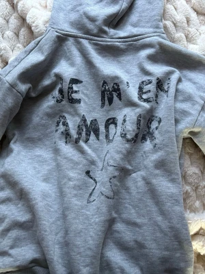 Grå hoodie med tryck 'Je m'en amour' - Säljer en grå hoodie med svart texttryck 'Je m'en amour' och en stjärna på ryggen. Hoodien har huva och är från Gina Young . Tröjan har andvändits X antal gånger men funkar fortfarande 