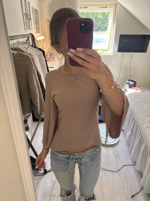 Beige ribbad topp med knytband - Säljer en beige ribbad topp med vida trekvartsärmar och knytband i midjan. Tröjan har en figurnära passform och är tillverkad i ett mjukt, stretchigt material. Perfekt att styla med jeans eller kjol för en trendig look.