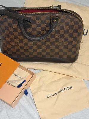 Authentic Louis Vuitton Alma handväska - Handväska från Louis Vuitton i klassiskt brunt damier-mönster. Väskan har allt inkluderat förutom boxen. Äktighetsbevis och kvitton finns med. Smått slitage, men väldigt omhändertagen. Pris går inte att diskuteras, då den säljs mindre än vad marknaden för just denna väska med ett sådant bra skick och inkluderande tillbehör säljs för. 