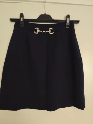 Mörkblå kjol med metallspänne - Stilren mörkblå kjol med ett dekorativt metallspänne framtill i midjan. Kjolen har en rak, klassisk form och är perfekt för dig som gillar en enkel men elegant look. Materialet känns mjukt och har en lätt struktur.