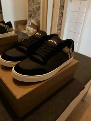 Svarta Burberry sneakers med rutdetalj - Snygga svarta sneakers från Burberry med klassisk beige och svart rutdetalj vid hälen. Skorna har vita sulor, svarta skosnören och en stilren design i läder och mocka. Perfekta för dig som vill ha en exklusiv touch på din outfit.
