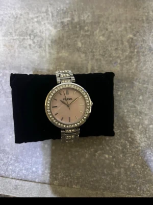 Silverfärgad klocka från Fossil - Stilren klocka från Fossil med silverfärgat armband och boett. Urtavlan är ljusrosa och omges av gnistrande stenar runt hela kanten. Armbandet har också stendetaljer som ger extra bling. Perfekt accessoar för dig som gillar en elegant look.