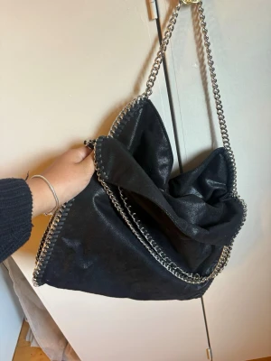 Handväska från Gina tricot  - Snygg svart axelväska i mjukt material med silverfärgade kedjor längs kanterna och som axelrem. Väskan har en slouchy form och ger en cool vibe till din outfit. Perfekt för dig som gillar statement-accessoarer och vill ha plats för allt du behöver. Knappt använd så i nyskick 