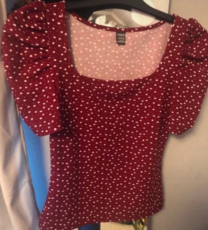 Vinröd topp med puffärm och hjärtan - Supersöt vinröd topp med vita hjärtan, fyrkantig ringning och puffiga korta ärmar. Materialet är stretchigt och mjukt, perfekt för en trendig look. Passar dig som gillar romantiska detaljer och vill sticka ut lite extra.