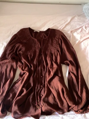 Brun boho-blus med pärldetaljer - Säljer en brun boho-inspirerad blus med långa ärmar och fina pärldetaljer i rött och gult vid knytbanden. Blusen har smockade partier upptill och knappar framtill. Luftigt material och avslappnad passform.