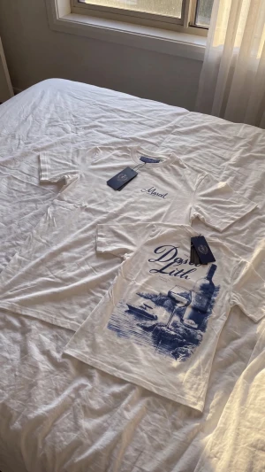 vita t-shirts med blått tryck - Säljer två vita t-shirts med blått tryck. Ena har texten 'Marcel' på bröstet, den andra har ett stort motiv med vinflaska, glas och texten 'Dess Luth'. Båda har rund hals och korta ärmar. Perfekta för en avslappnad stil.