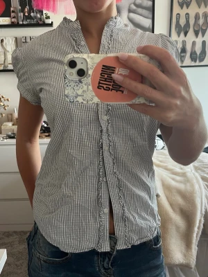 Randig kortärmad skjorta/blus med volang - Säljer en söt kortärmad skjorta med smala blå och vita ränder. Skjortan har volangdetaljer längs knappraden och en liten ståkrage. Tillverkad i tunn bomull, perfekt för varmare dagar. Passformen är normal och skjortan har knappar framtill.