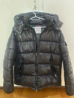 Svart dunjacka från Moncler med huva - Snygg svart dunjacka från Moncler med glansig finish och avtagbar huva. Jackan har två dragkedjeförsedda fickor framtill och en hög krage. Perfekt för kalla vinterdagar och riktigt trendig look.