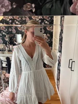 Vit kort klänning från Zara - Söt vit kort klänning från Zara med v-ringning, spetsdetaljer och knappar framtill. Klänningen har långärmade ärmar och volang nedtill, samt ett lätt och luftigt tyg som passar perfekt till varma dagar.🌸