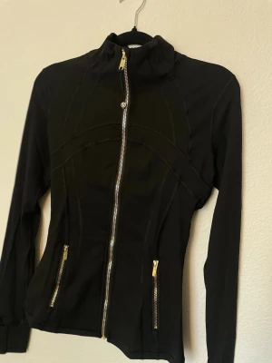 Lululemon xs/ 6 - Snygg svart vindjacka med figurnära passform och guldiga dragkedjor framtill och på fickorna. Jackan har hög krage och långa ärmar, perfekt för en sportig och stilren look. Materialet känns lätt och smidigt, vilket gör den enkel att bära till vardags.