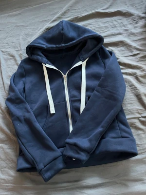 Mörkblå zip hoodie med vita snören - Mysig mörkblå hoodie med dragkedja framtill och vita snören i huvan. Klassisk känguruficka och ribbade muddar vid ärmslut och nederkant. Perfekt för chill dagar eller när du vill ha en enkel och stilren look. Materialet känns mjukt och skönt mot huden.
