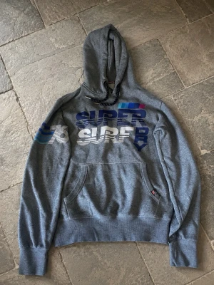Grå Superdry hoodie med tryck - Snygg grå hoodie från Superdry med stort färgglatt tryck på bröstet där det står 'Super Surf Dry' och blåa ränder på ena ärmen. Klassisk känguruficka framtill och justerbar huva med snören. Perfekt för en avslappnad streetwear-look. Som ny i skicket.