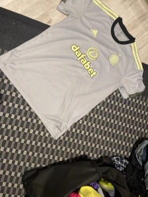 Grå Celtic FC matchtröja Adidas - Snygg grå matchtröja från Celtic FC med gula detaljer och svarta inslag. Adidas-logga på bröstet, klubbmärke och sponsortryck fram och bak. Tillverkad i lätt och ventilerande funktionsmaterial, perfekt för träning eller match. Can negotatiate for lower price