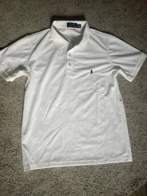 Vit pikétröja från Polo Ralph Lauren - Klassisk vit pikétröja från Polo Ralph Lauren med blå logga broderad på bröstet. Tröjan har korta ärmar, krage och knappar framtill. Tillverkad i bomull för en skön och luftig känsla.