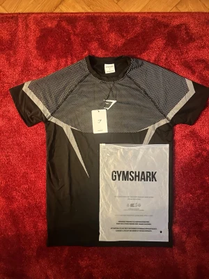Svart/vit Gymshark träningströja S - Säljer en svart träningströja från Gymshark i storlek S. Tröjan har gråa och vita detaljer över bröst och axlar samt Gymshark-logga framtill. Tillverkad i ett lätt och snabbtorkande material, perfekt för gymmet eller löpning.