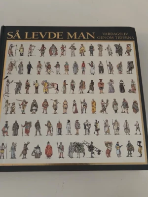 Så levde man - vardagsliv genom tiderna - En illustrerad bok med titeln 'Så levde man - vardagsliv genom tiderna'. Omslaget är svart med guldfärgad text och visar många små tecknade figurer i olika historiska kläder. Perfekt för dig som gillar historia och kultur.