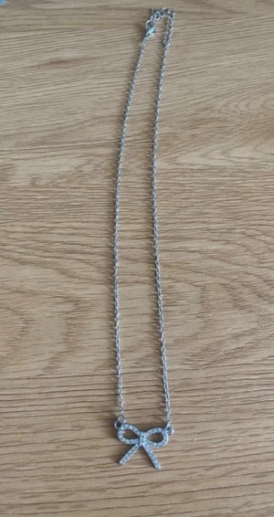 Silverfärgat halsband med rosett - Snyggt halsband i silverfärg med ett hänge format som en rosett, dekorerat med små gnistrande stenar. Kedjan är tunn och elegant, perfekt för att ge en söt touch till din outfit. Passar dig som gillar detaljer och glitter.