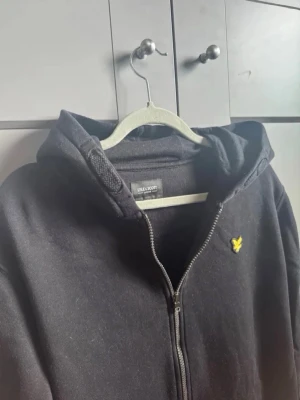 Lyle & Scott zip - Svart hoodie från Lyle & Scott med dragkedja framtill och klassisk huva. Bröstet pryds av den ikoniska gula loggan. Hoodien har två fickor framtill och är tillverkad i mjukt bomullsmaterial. Perfekt för en avslappnad och stilren look. XL i barnstorlek från kidsbrandstore(sitter som S)